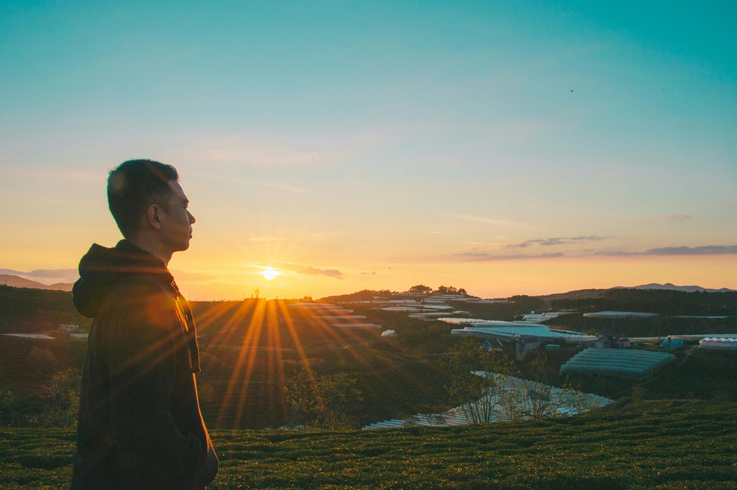 Silhouette of a man in a hoodie admiring a sunrise over rural landscapes.